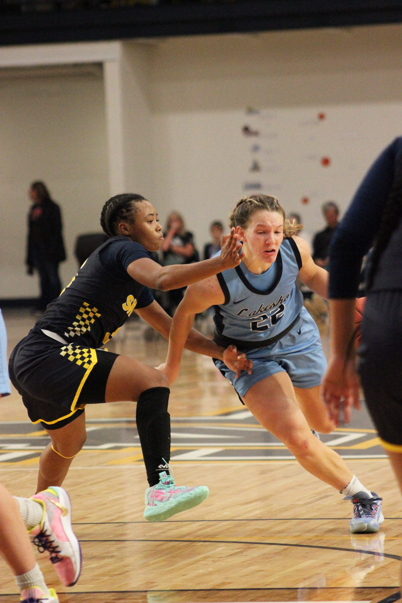 Two female basketball players during a game. Their jerseys read "lakers" and "skippers". The players with a "skippers" jersey is trying to block the player with a "lakers" jersey from running past. 