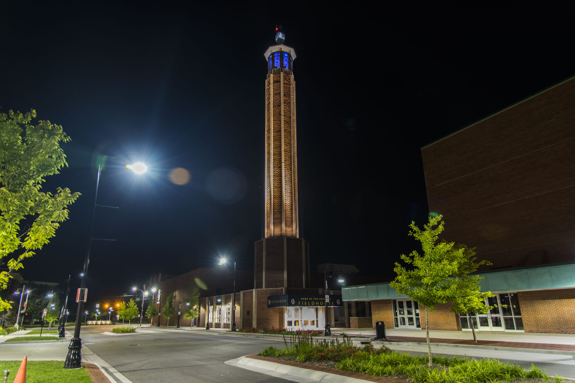 A large building on a city street at night. The building has a banner above the door that reads "fieldhouse". The building has a high tower and the top is illuminated by blue lights. 