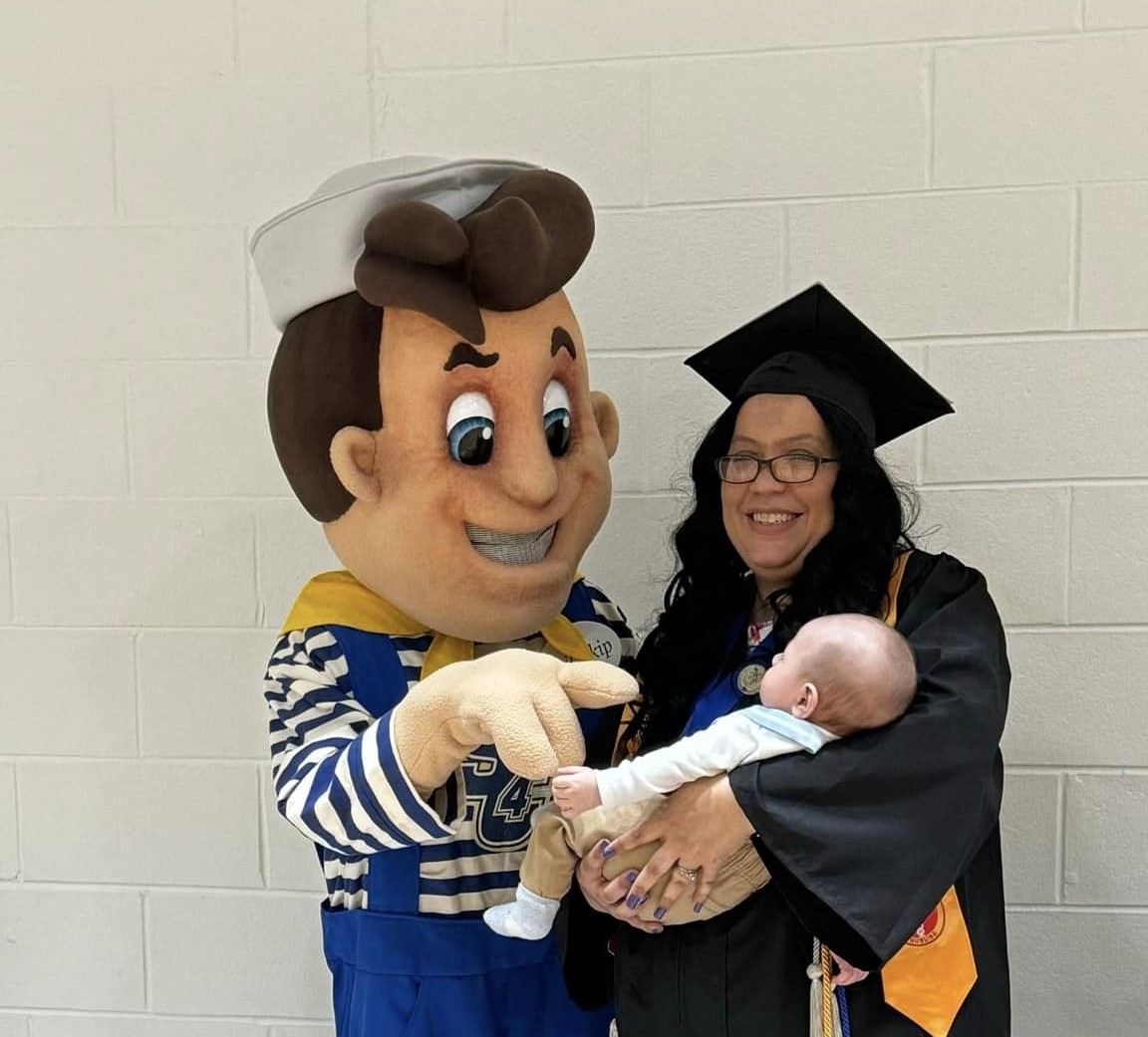 Lil Skip mascot standing next to a woman in graduation robes. The woman is holding a baby in her arms. 